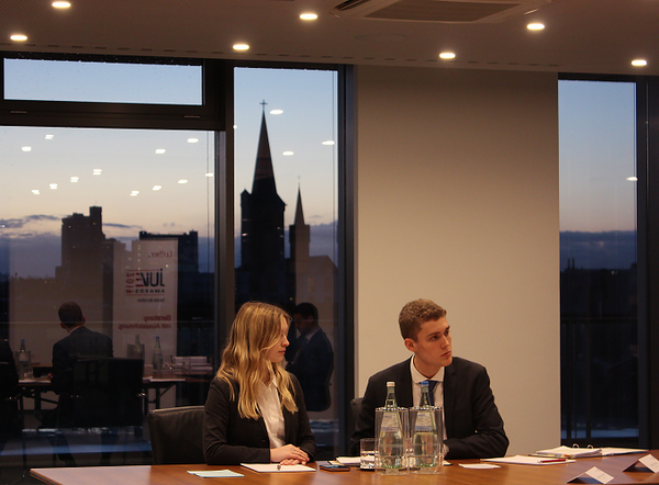 Zwei Personen in formeller Kleidung sitzen an einem Konferenztisch. Die Frau trägt eine schwarze Jacke und hat lange blonde Haare. Der Mann hat kurzes, helles Haar. Im Hintergrund sind Stadtgebäude und ein historischer Kirchturm bei Sonnenuntergang sichtbar.