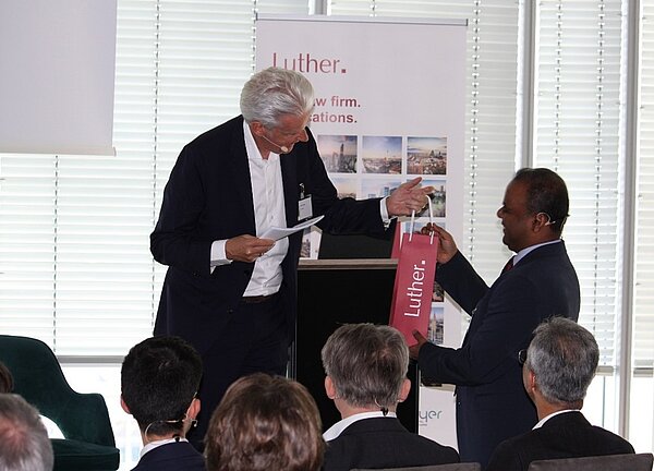 Zwei Männer stehen auf einer Veranstaltung, während einer einen Geschenktüte mit dem Schriftzug "Luther" überreicht. Der Mann auf der linken Seite hält ein Blatt Papier in der Hand. Im Hintergrund sind Teilnehmer, die dem Geschehen aufmerksam folgen.