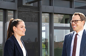 Zwei Personen stehen im Freien, vor einem modernen Gebäude. Eine Frau mit langen Haaren trägt einen blauen Blazer und lächelt, während ein Mann in einem Anzug und einer Brille ebenfalls lächelt und sie ansieht. Beide wirken freundlich und in einem Gespräch vertieft.
