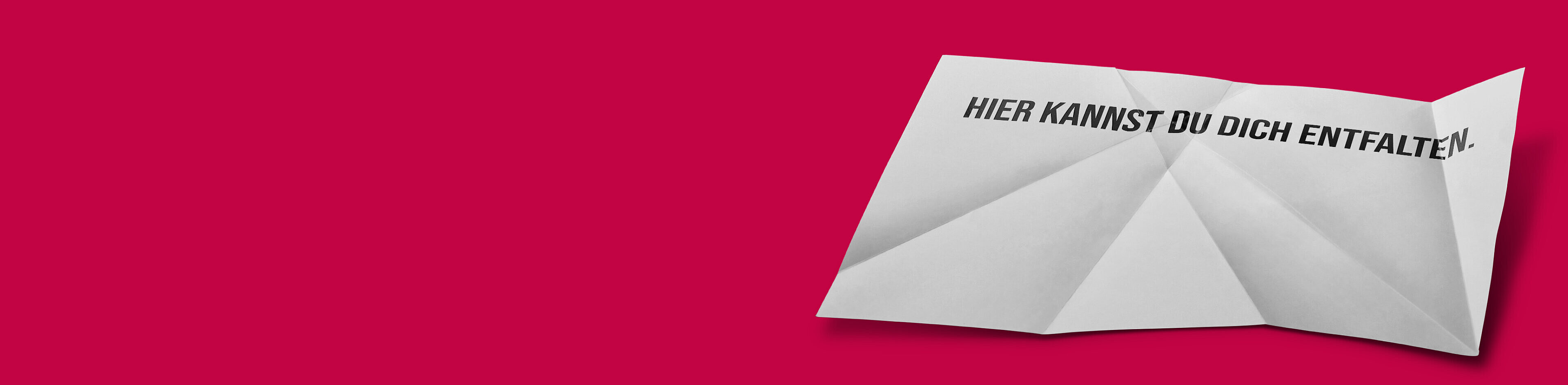 Ein zusammengefaltetes, weißes Blatt Papier mit der Aufschrift „HIER KANNST DU DICH ENTFALTEN.“ auf einem roten Hintergrund. Das Bild vermittelt eine Botschaft der Entfaltung und Möglichkeiten.