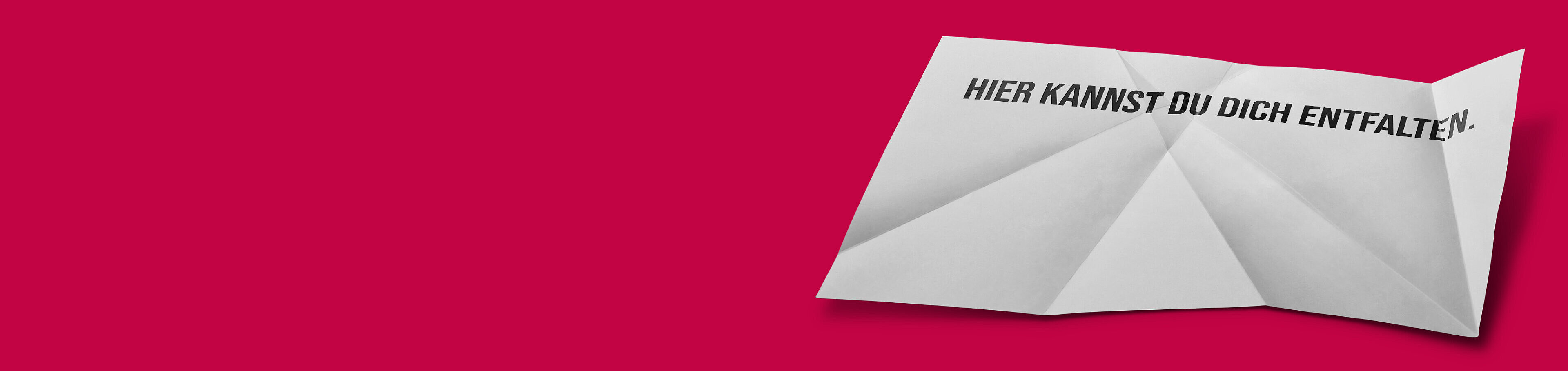 Ein zusammengefaltetes, weißes Blatt Papier mit der Aufschrift „HIER KANNST DU DICH ENTFALTEN.“ auf einem roten Hintergrund. Das Bild vermittelt eine Botschaft der Entfaltung und Möglichkeiten.