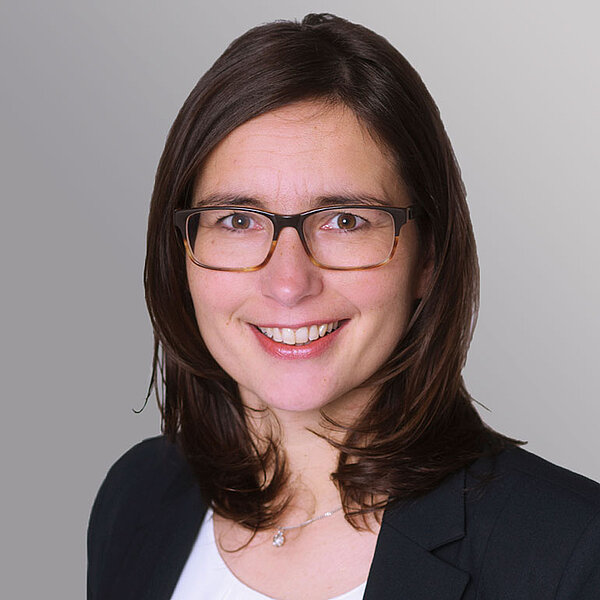 Ein Porträt einer lächelnden Frau mit langen, dunklen Haaren und Brille. Sie trägt eine dunkle Blazerjacke über einem weißen Oberteil. Der Hintergrund ist grau, was einen professionellen Eindruck vermittelt.