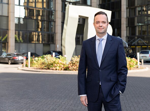 Ein Mann in einem dunklen Anzug und hellblauen Krawatte steht lächelnd im Freien vor einem modernen Gebäude mit großen Fenstern und einer skulpturalen Installation. Die Umgebung ist urban mit einigen Fahrzeugen im Hintergrund.