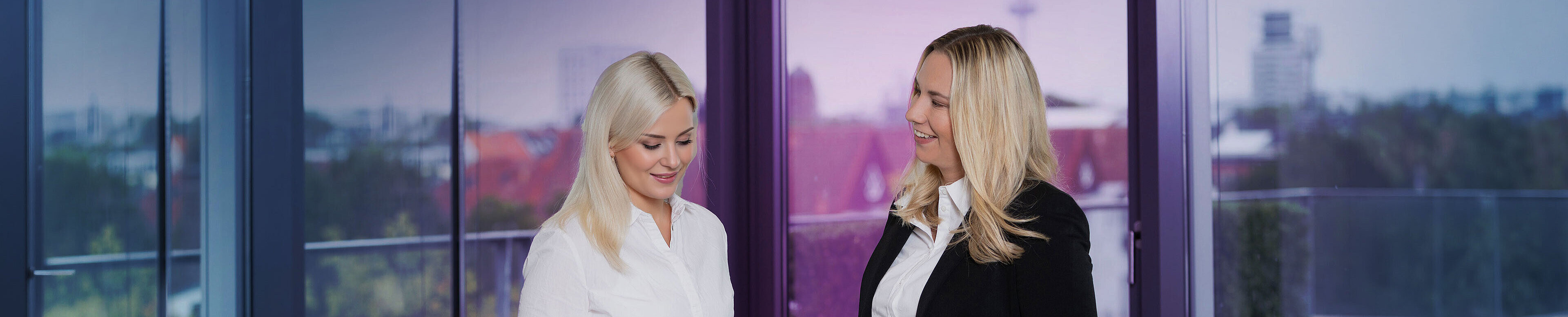 Zwei Frauen in formeller Kleidung stehen in einem modernen Büro mit großen Fenstern. Eine Frau hält ein Tablet und zeigt es der anderen, während sie miteinander sprechen. Im Hintergrund ist eine Stadtansicht sichtbar.