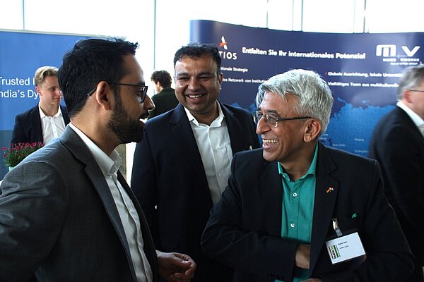 Zwei Männer in Anzügen unterhalten sich lächelnd miteinander. Der Mann links trägt eine Brille und hat einen Bart, während der andere graue Haare und ein grünes Hemd trägt. Im Hintergrund sind verschwommene Silhouetten anderer Personen und ein Werbebanner zu sehen.