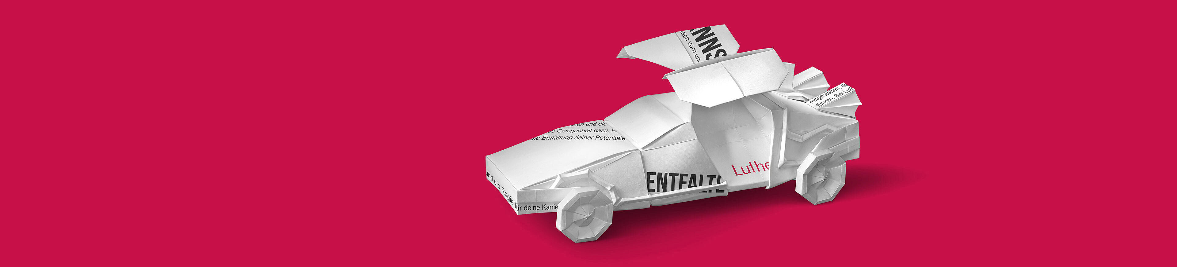 Ein origamiartiges Auto aus weißem Papier steht auf einem roten Hintergrund. Es sind Texte und Hinweise auf diesem Auto zu sehen, was auf eine kreative Präsentation oder eine Botschaft hinweist.