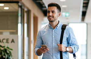 Ein junger Mann mit dunklen Haaren trägt ein hellblaues Hemd und eine Schultertasche. Er hält ein Smartphone in der Hand und lächelt, während er durch einen modernen Flur geht. Im Hintergrund sind Pflanzen und Büroelemente zu sehen.