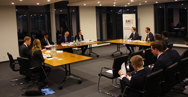 Eine Gruppe von Personen sitzt an einem langen Tisch in einem modern gestalteten Raum. Sie diskutieren und präsentieren in einem formellen Rahmen. Wasserkrüge und Gläser sind auf dem Tisch platziert, während ein Banner im Hintergrund sichtbar ist. Die Atmosphäre wirkt professionell und konzentriert.