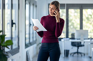 Eine Frau spricht am Telefon, während sie eine Papierseite in der Hand hält. Sie trägt einen roten Pullover und steht in einem hellen, modernen Büro mit großen Fenstern. Im Hintergrund sind Schreibtische und Bürostühle sichtbar.
