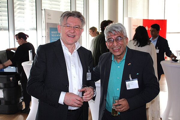 Zwei Männer stehen lächelnd nebeneinander auf einer Veranstaltung. Der Mann auf der linken Seite trägt ein schwarzes Sakko und eine weiße Bluse, während der Mann rechts ein dunkles Jackett und ein türkisfarbenes Hemd anhat. Beide halten Gläser in der Hand.