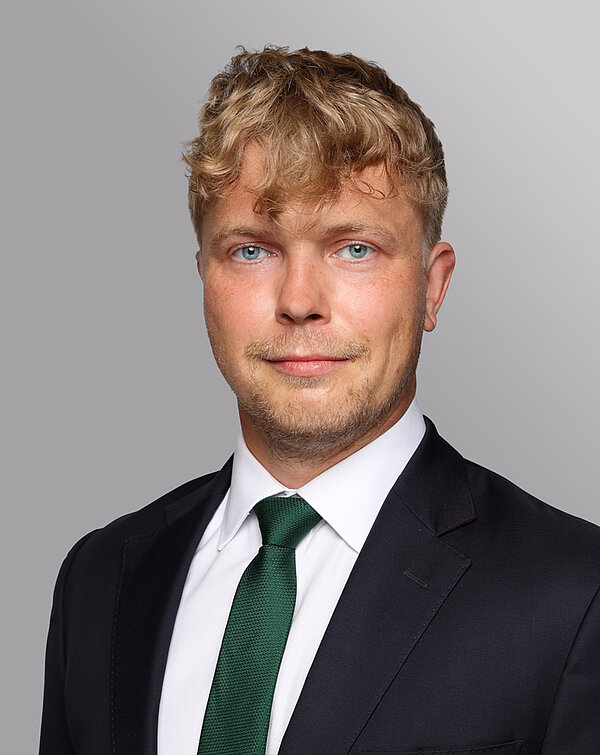 Ein junger Mann mit lockigem, blondem Haar lächelt in die Kamera. Er trägt einen schwarzen Anzug mit weißem Hemd und einer grünen Krawatte. Der Hintergrund ist grau.