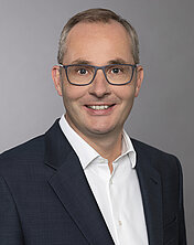 Porträt eines lächelnden Mannes mit kurzen, grauen Haaren und Brille. Er trägt einen dunklen Blazer über einem weißen Hemd. Der Hintergrund ist grau und neutral, um den Fokus auf das Gesicht zu legen.