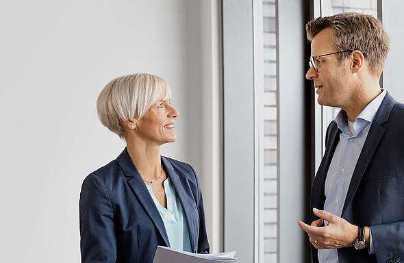 Zwei Personen in formeller Kleidung stehen in einem modernen Büro nebeneinander. Sie unterhalten sich, während die Frau lächelt und ein paar Blätter in der Hand hält. Im Hintergrund ist ein Fenster mit Blick auf eine Stadtlandschaft.
