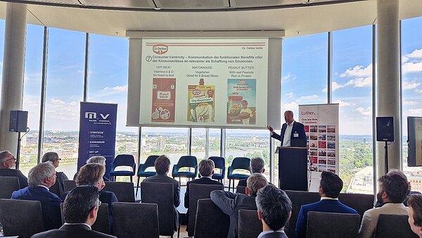 Ein Vortragender präsentiert Informationen über Lebensmittelmarken wie Amul, Funfoods und Real Peanut Butter. Im Hintergrund ist ein grafisches Element zu sehen, das die Kommunikation funktionaler Vorteile für Konsumenten betont. Das Publikum im Vordergrund besteht aus mehreren Anwesenden, die ihm zuhören.