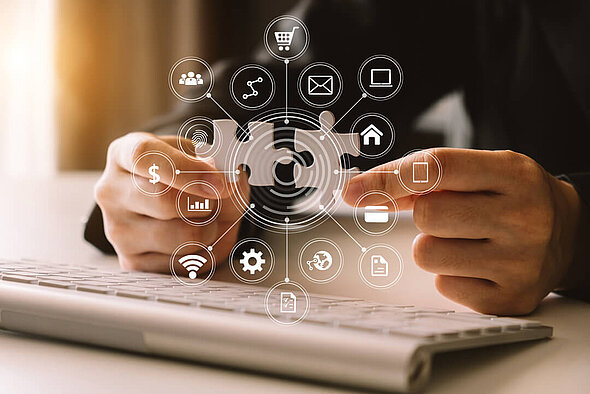 Eine Person sitzt an einem Computer und zeigt mit zwei Fingern auf ein Puzzle-Teil. Um das Puzzle-Teil sind verschiedene Symbole angeordnet, die digitale Dienstleistungen und Kommunikation darstellen, wie z.B. E-Mail, Shopping und Datenanalyse, was auf eine Verbindung von Ideen hinweist.