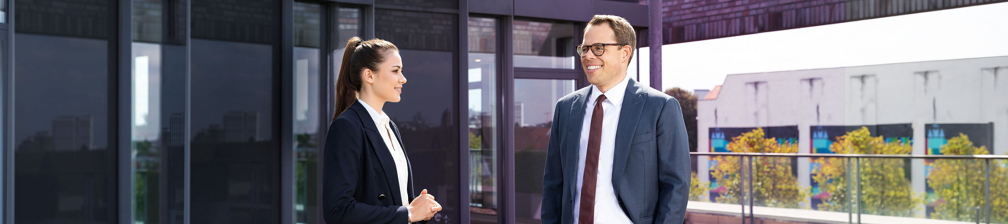 Zwei Personen in Businesskleidung stehen auf einer modernen Terrasse. Die Frau hat lange Haare und trägt einen dunklen Anzug, während der Mann eine Brille und einen grauen Anzug mit einer roten Krawatte trägt. Im Hintergrund sind moderne Gebäude und Bäume sichtbar.
