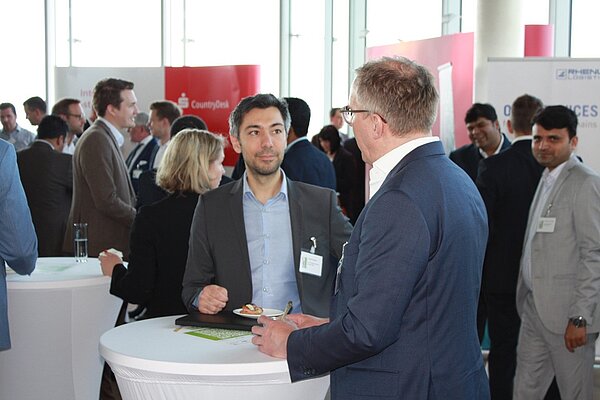 Eine Gruppe von Menschen steht in einem modernen Eventraum und unterhält sich. Zwei Männer im Vordergrund sind in ein Gespräch vertieft. Einer von ihnen hält einen kleinen Teller mit Essen, während der andere Notizen macht. Hintergrundaktivitäten zeigen Networking-Stimmung.