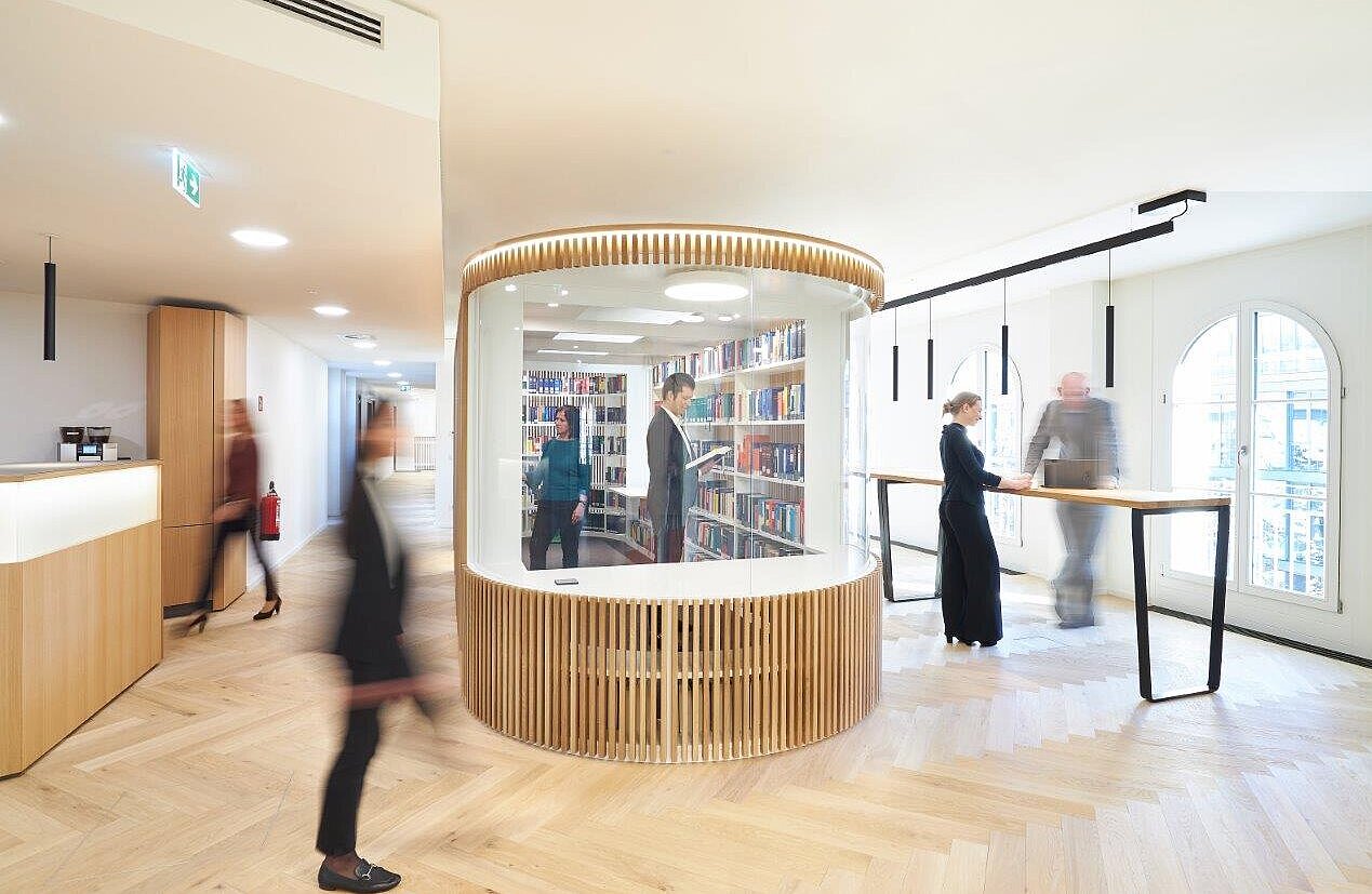 Das Bild zeigt einen modernen, hellen Raum mit Bücherregalen. In einem gläsernen Bereich stehen mehrere Personen, die Bücher lesen oder betrachten. Im Hintergrund sind weitere Personen sichtbar, die sich im Raum bewegen. Das Interieur ist minimalistisch und freundlich gestaltet.