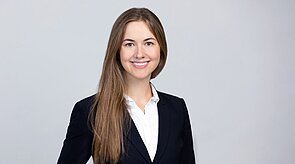 Eine lächelnde Frau mit langen, braunen Haaren trägt ein schwarzes Jacket und ein weißes Hemd. Hintergrund ist hellgrau. Sie steht aufrecht und präsentiert ein selbstbewusstes Auftreten.