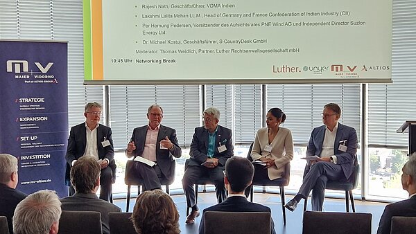 Eine Diskussionsrunde mit fünf Rednern, die auf Stühlen sitzen und über strategische Themen sprechen. Im Hintergrund sind Fenster mit Jalousien zu sehen. Auf einem Bildschirm im Hintergrund sind Namen und Funktionen der Teilnehmer sowie das Veranstaltungsthema angezeigt.