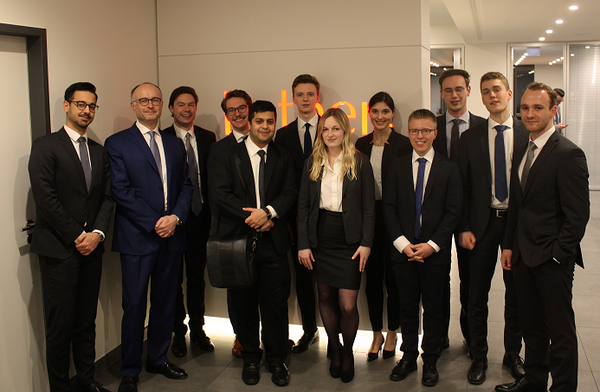 Eine Gruppe von elf Personen in formeller Kleidung steht vor einer Wand mit dem Schriftzug "Norton". Die Menschen lächeln und posieren in einer modernen Büroumgebung. Sie sind sowohl Männer als auch Frauen, die unterschiedliche kulturelle Hintergründe repräsentieren.