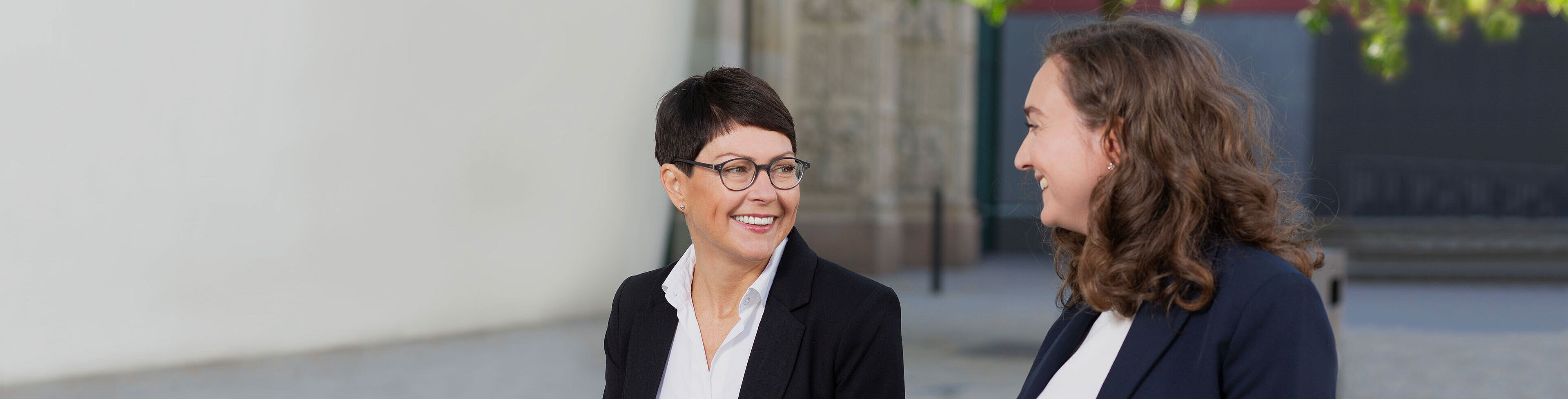 Zwei Frauen in Anzügen sitzen zusammen und lachen. Sie wirken freundlich und unterhalten sich angeregt. Im Hintergrund sind unscharfe Gebäudeteile und ein Baum sichtbar.