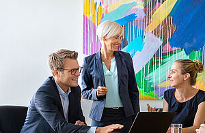 Drei Personen in formeller Kleidung unterhalten sich lebhaft in einem modernen Büro. Ein Mann sitzt vor einem Laptop, während eine Frau steht und spricht. Eine weitere Frau hört aufmerksam zu. Im Hintergrund ist ein farbenfrohes, abstraktes Kunstwerk zu sehen.