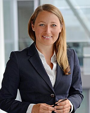 Eine lächelnde Frau steht im Vordergrund, trägt einen dunklen Anzug und ein weißes Hemd. Sie hält einen Stift in der Hand und wirkt professionell und freundlich. Der Hintergrund ist unscharf, mit Anzeichen eines modernen Büroumfelds.