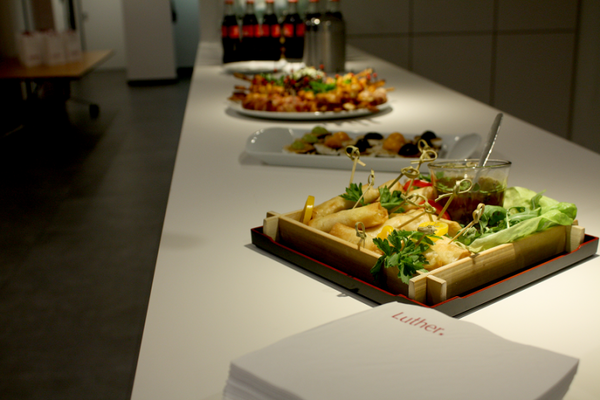 Ein langes Tischbuffet mit verschiedenen Speisen. Im Vordergrund ein Tablett mit gefüllten Teigtaschen, frischen Kräutern und Salat. Im Hintergrund sind mehrere Platten mit Fingerfood und Getränken sichtbar. Ein weißes Serviettenpaket mit dem Schriftzug „Luther“ liegt auf dem Tisch.