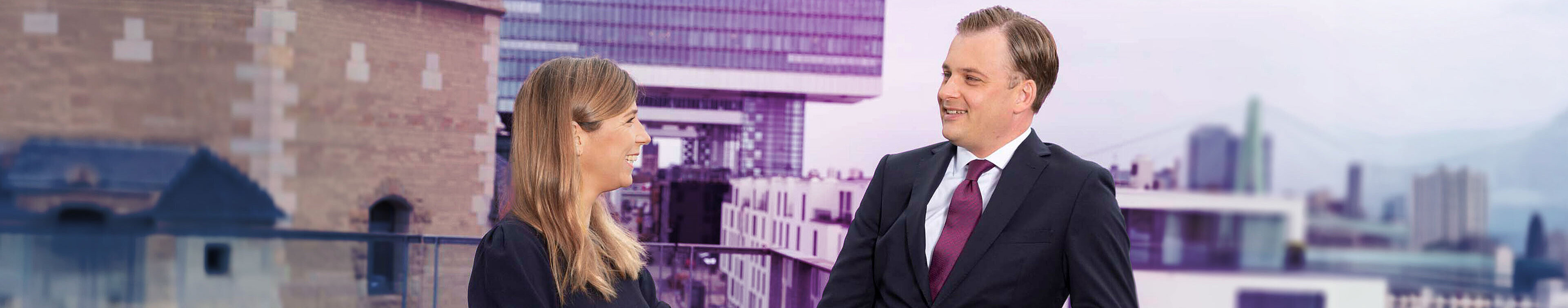 Zwei Personen stehen auf einer Dachterrasse und unterhalten sich. Im Hintergrund sind moderne Gebäude und Stadtlandschaft sichtbar. Die Frau trägt ein schwarzes Kleid, der Mann einen Anzug mit Krawatte. Die Szene vermittelt eine professionelle Atmosphäre.
