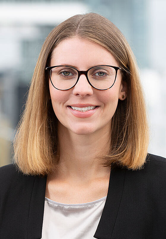 Porträt einer Frau mit langen, hellbraunen Haaren und einer Brille. Sie trägt einen schwarzen Blazer und lächelt freundlich. Der Hintergrund ist unscharf und zeigt eine moderne Gebäudeansicht.