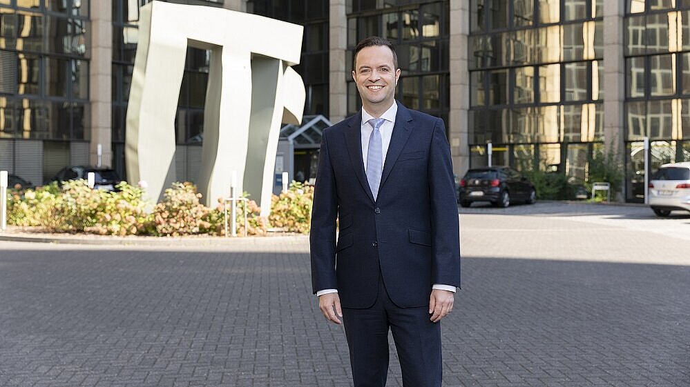 Ein Mann in einem Anzug steht lächelnd vor einem modernen Gebäude mit vielen Fenstern. Hinter ihm ist eine große, abstrakte Skulptur zu sehen. Die Umgebung ist gepflegt mit einigen Pflanzen und parkenden Autos.