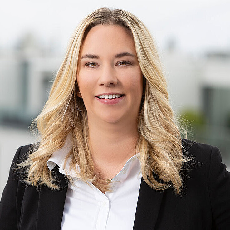 Ein Porträt einer lächelnden Frau mit blonden, lockigen Haaren, die ein schwarzes Blazerjackett und ein weißes Hemd trägt. Hintergrund zeigt unscharfe Gebäudestrukturen und einen bewölkten Himmel. Die Frau steht auf einer Terrasse und wirkt freundlich und professionell.