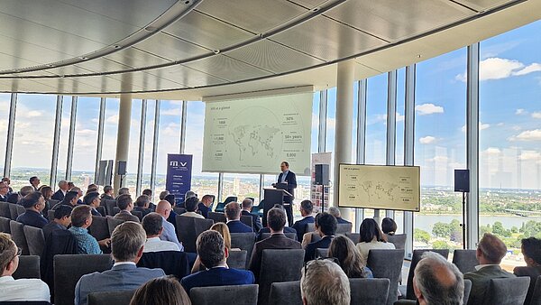 Ein Mann steht an einem Podium und präsentiert Informationen auf einer großen Leinwand in einem modernen Veranstaltungsraum. Im Hintergrund sind Mitarbeiter und Gäste im Publikum zu sehen, die aufmerksam zuhören. Durch große Fenster ist eine Aussicht auf die Stadt sichtbar.
