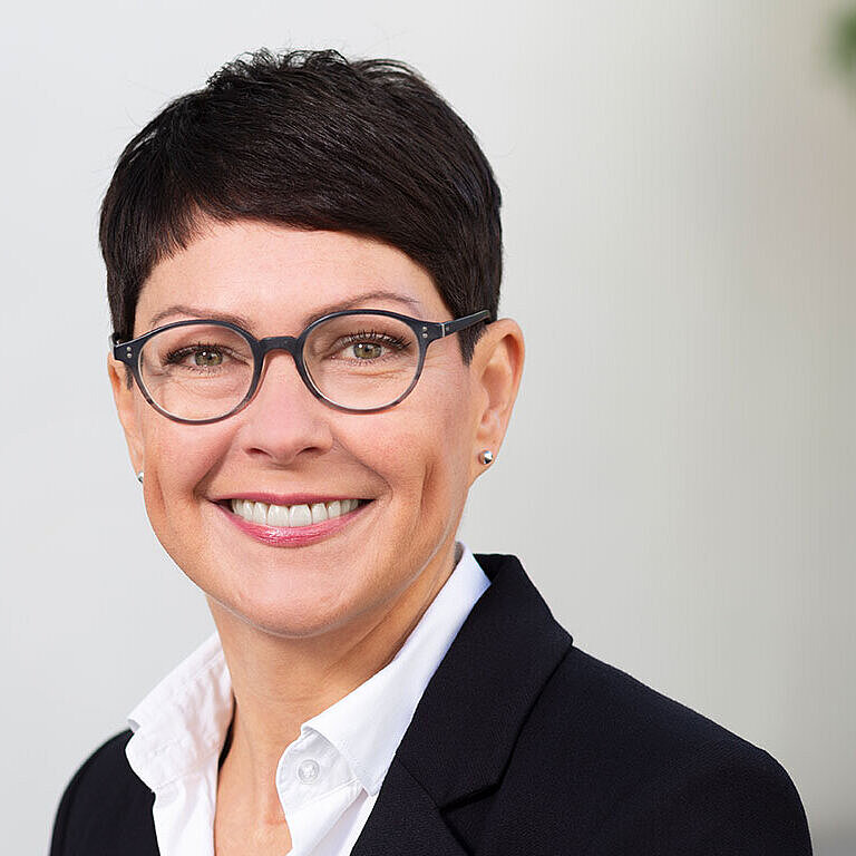 Porträt einer lächelnden Frau mit kurzem, dunklem Haar und Brille, die einen schwarzen Blazer und ein weißes Hemd trägt. Der Hintergrund ist hell und neutral gehalten. Die Frau wirkt freundlich und professionell.