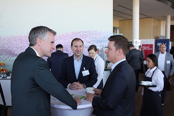 Eine Gruppe von vier Personen unterhält sich an einem Stehtisch bei einer Veranstaltung. Zwei Männer sprechen intensiv miteinander, während ein dritter Mann aufmerksam zuhört. Im Hintergrund sind weitere Teilnehmer und ein Buffet sichtbar. Eine Bedienung trägt ein Tablett mit Getränken.