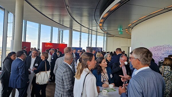 Eine Gruppe von Menschen steht in einem modernen Raum mit großen Fenstern. Sie unterhalten sich und halten Getränke in der Hand. Im Hintergrund sind Wandverkleidungen und Informationsstände zu sehen, die eine geschäftliche Veranstaltung andeuten.
