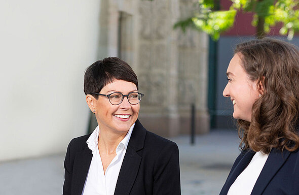 Zwei Frauen in formeller Kleidung stehen im Freien und lächeln einander freundlich zu. Die Frau links hat kurze, dunkle Haare und trägt eine Brille, während die Frau rechts lockige, mittellange Haare hat. Im Hintergrund sind verschwommene architektonische Elemente zu erkennen.