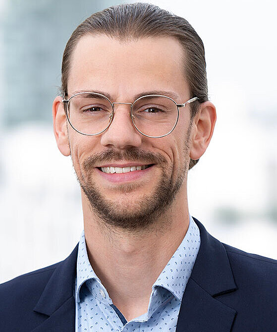Ein junger Mann mit Brille lächelt freundlich in die Kamera. Er trägt einen dunkelblauen Anzug und ein hellblaues Hemd mit Punktmuster. Der Hintergrund ist unscharf und zeigt einen städtischen Ausblick.