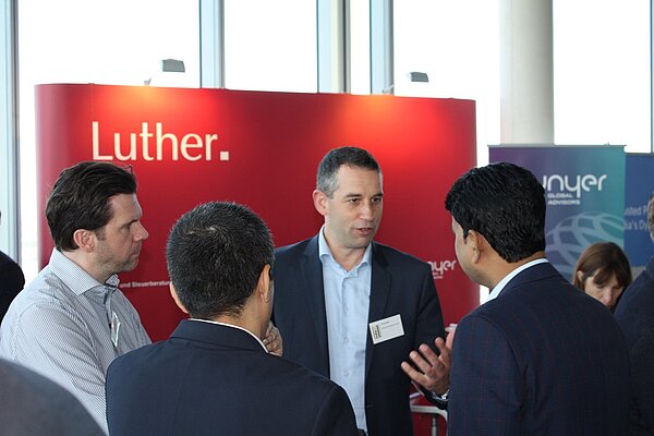 Eine Gruppe von vier Männern steht an einem Stand und unterhält sich. Einer der Männer gestikuliert, während er spricht. Im Hintergrund sind zwei farbige Banner zu sehen, auf einem steht der Name "Luther". Die Szene vermittelt ein Gefühl von Networking und Austausch.