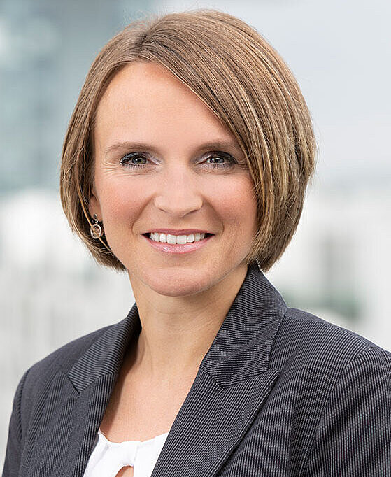 Porträt einer Frau mit kurzem, braunem Haar. Sie trägt ein graues Blazer und lächelt, während sie direkt in die Kamera schaut. Der Hintergrund ist unscharf und zeigt eine helle, moderne Umgebung.