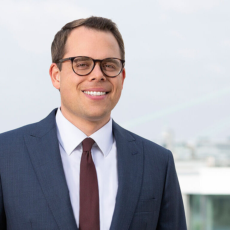 Ein junger Mann mit Brille und einem dunkelblauen Anzug lächelt freundlich. Er steht vor einem verschwommenen, urbanen Hintergrund mit modernen Gebäuden. Sein lässiger, professioneller Look vermittelt Selbstbewusstsein und Freundlichkeit.