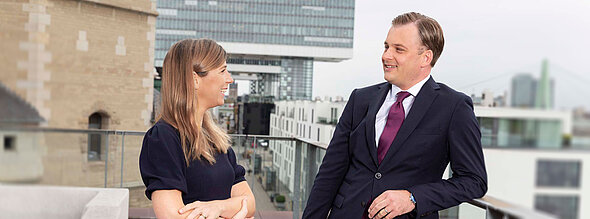 Zwei beruflich gekleidete Personen stehen auf einer Terrasse und unterhalten sich. Die Frau trägt ein dunkles Kleid, der Mann einen Anzug mit Krawatte. Im Hintergrund sind moderne Gebäude und eine Brücke zu sehen, die eine städtische Umgebung darstellt.