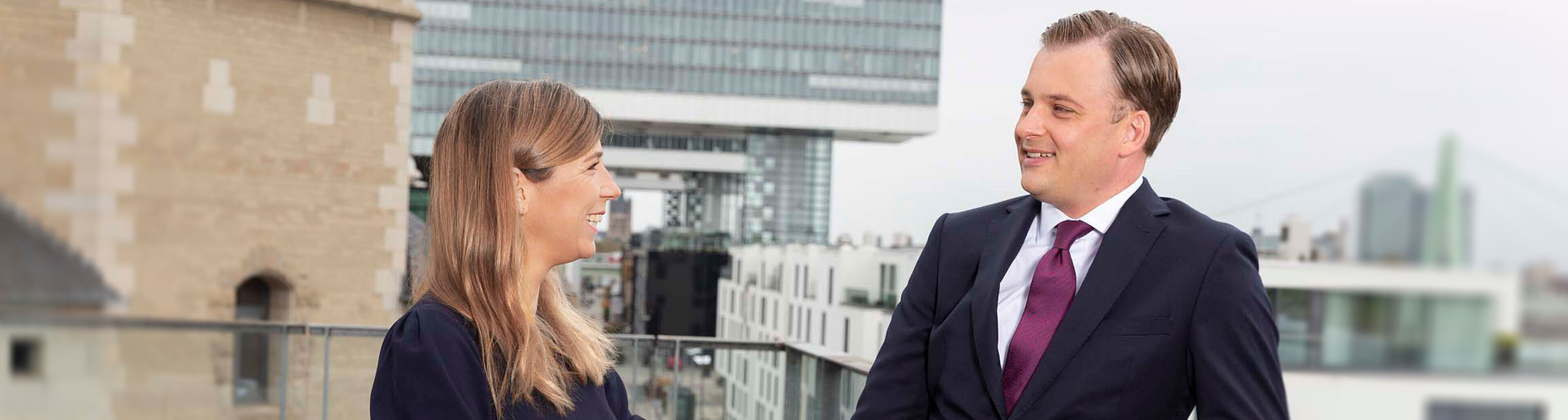 Zwei beruflich gekleidete Personen stehen auf einer Terrasse und unterhalten sich. Die Frau trägt ein dunkles Kleid, der Mann einen Anzug mit Krawatte. Im Hintergrund sind moderne Gebäude und eine Brücke zu sehen, die eine städtische Umgebung darstellt.