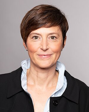 Porträt einer Frau mit kurzem, braunem Haar. Sie trägt einen schwarzen Blazer über einem hellblauen Hemd. Die Frau schaut freundlich und überzeugt in die Kamera, mit einem leichten Lächeln auf den Lippen. Der Hintergrund ist schlicht und hellgrau.