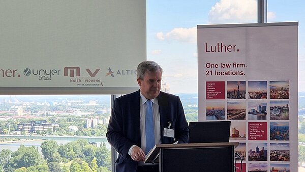 Ein Mann steht an einem Podium und spricht. Im Hintergrund ist ein Banner der Kanzlei Luther mit dem Slogan „One law firm. 21 locations.“ und Auflistungen von Städten, in denen die Kanzlei vertreten ist. Die Sicht auf eine grüne Landschaft und den Himmel ist zu erkennen.