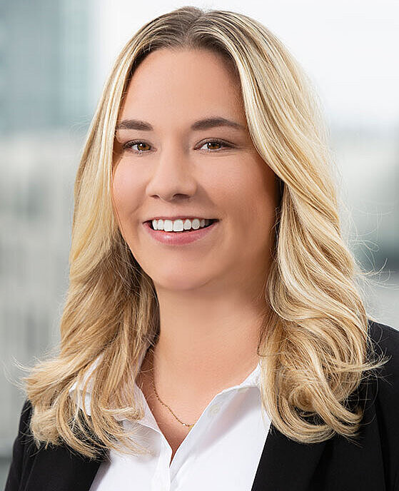 Eine lächelnde Person mit langen, blonden Haaren trägt ein schwarzes Blazerjackett und ein weißes Hemd. Der Hintergrund ist unscharf und zeigt eine moderne Büroumgebung.