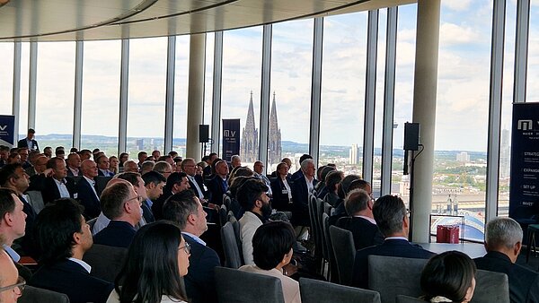 Eine Gruppe von Menschen sitzt in einem modernen Konferenzraum mit großen Fenstern. Im Hintergrund sind die Türme eines berühmten Gebäudes sichtbar. Die Teilnehmer hören aufmerksam zu, während eine Präsentation stattfindet.