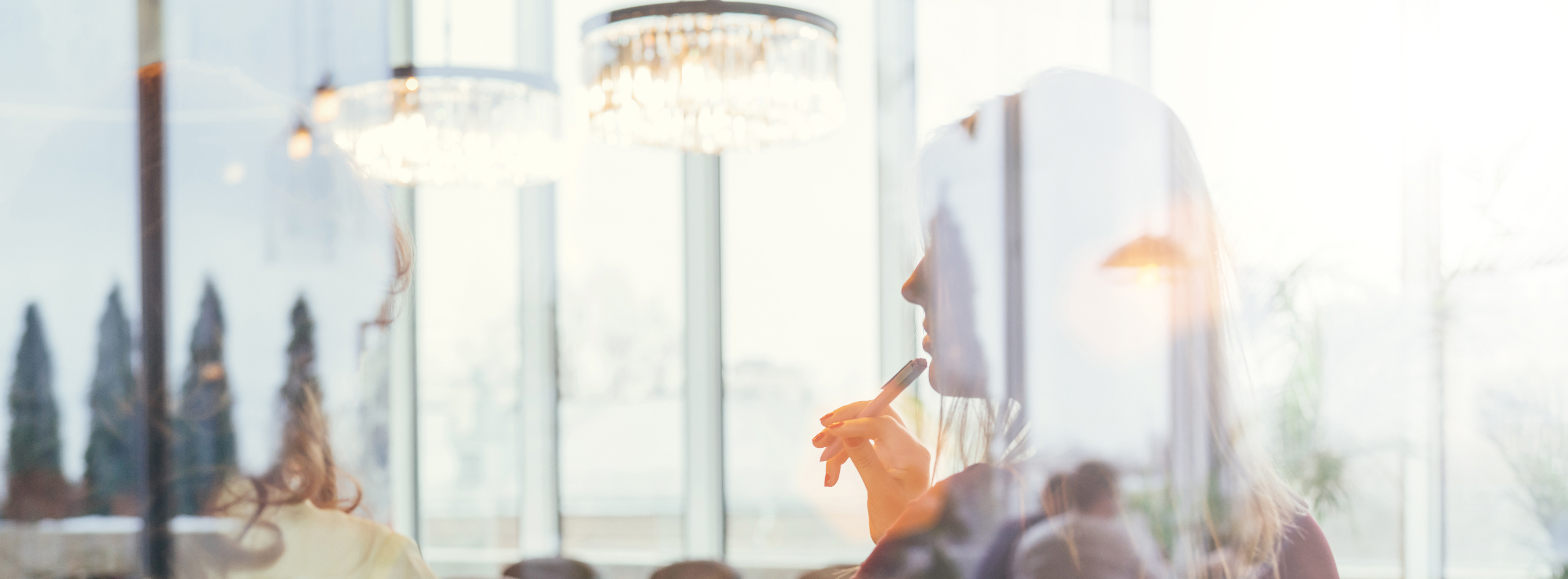 Eine Frau sitzt in einem hellen Raum und hält einen Stift an ihren Mund. Im Hintergrund sind große Fenster und elegante Leuchten sichtbar. Die Atmosphäre wirkt kreativ und nachdenklich.