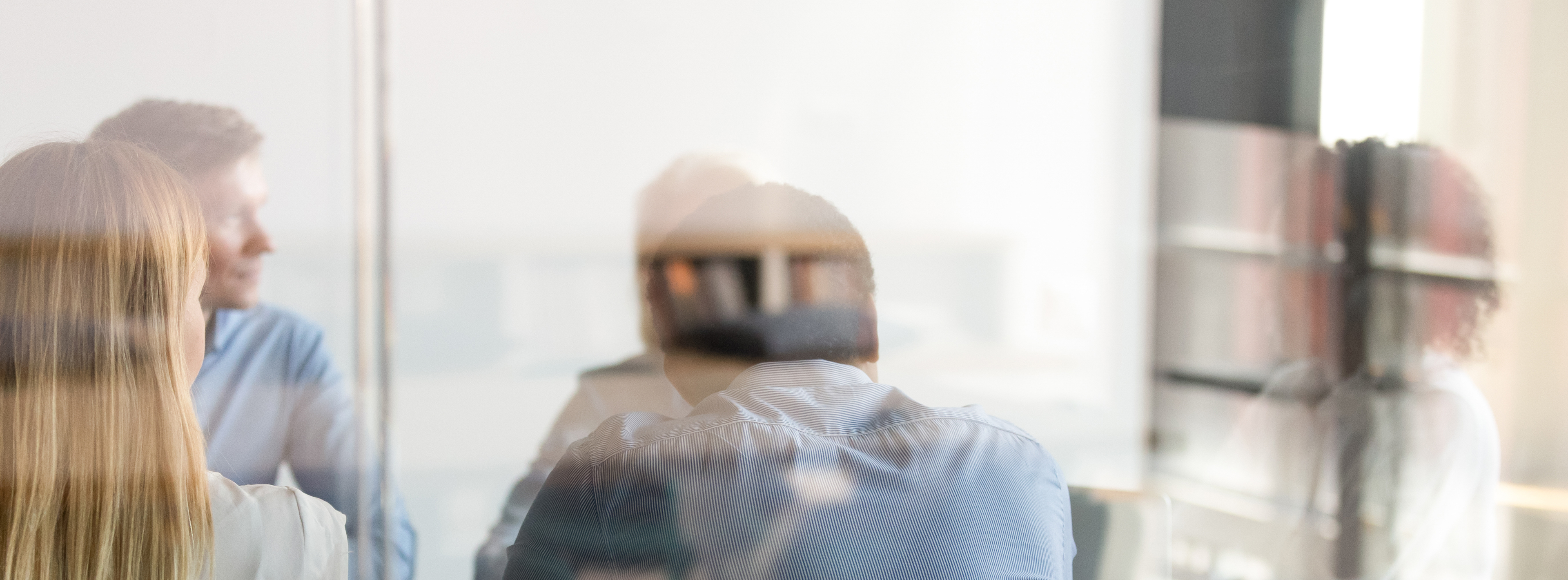 Eine Gruppe von mehreren Menschen in einem Besprechungsraum, die in unterschiedlicher Richtung sitzen. Die Atmosphäre wirkt entspannt, während sie miteinander kommunizieren. Die Bildkomposition ist leicht unscharf, was die Gesichter und Details der Personen nur teilweise sichtbar macht.